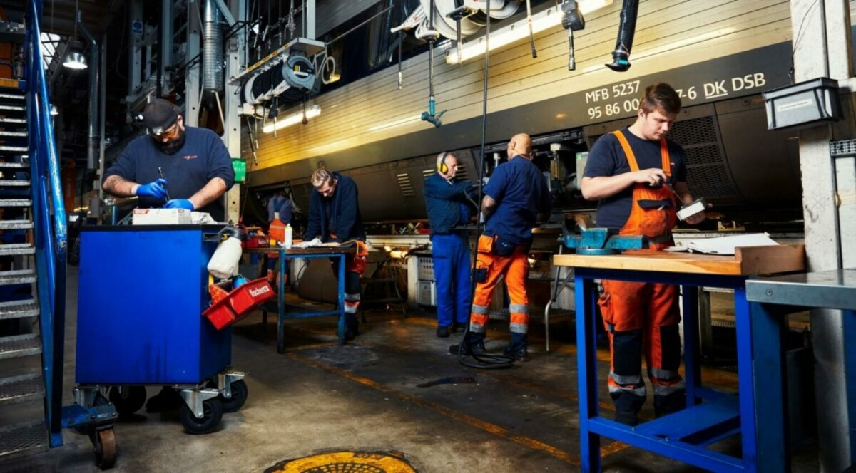 DSB-værksted med fem håndværkere i blå t-shirts og orange smækbukser reparerer ting. I baggrunden et hvidt og sort IC3-tog