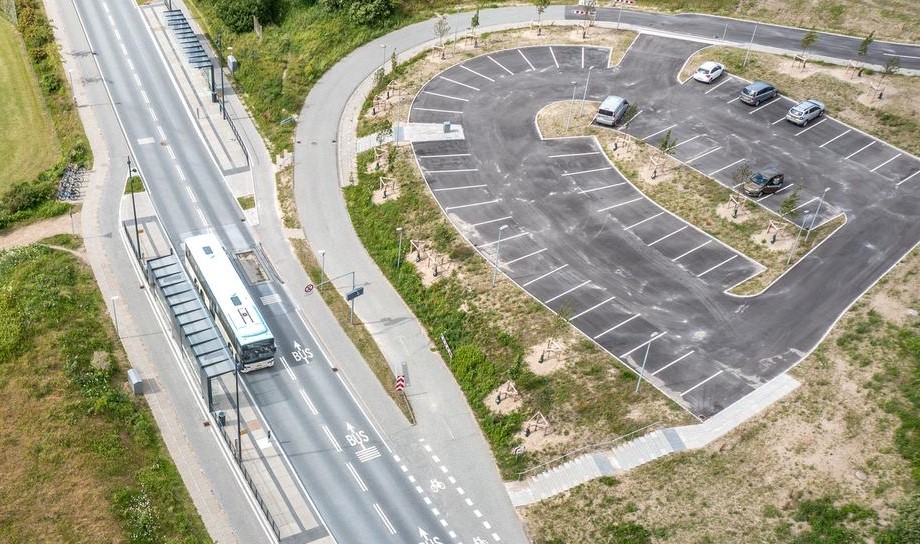 Dronefoto af knudepunktet. Der holder fem biler parkeret på den ovale p-plads. En Plusbus holder ved bosstoppestedet.
