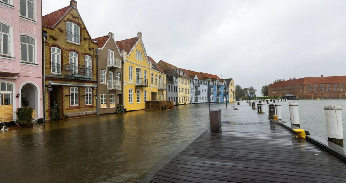 Lyserøde, gule, blå og røde huse på række på Sønderborg Havn. Gaden er dækket af vand. Der er grønne sandsække ved dørene. Gaden går i et med vandet i sundet.