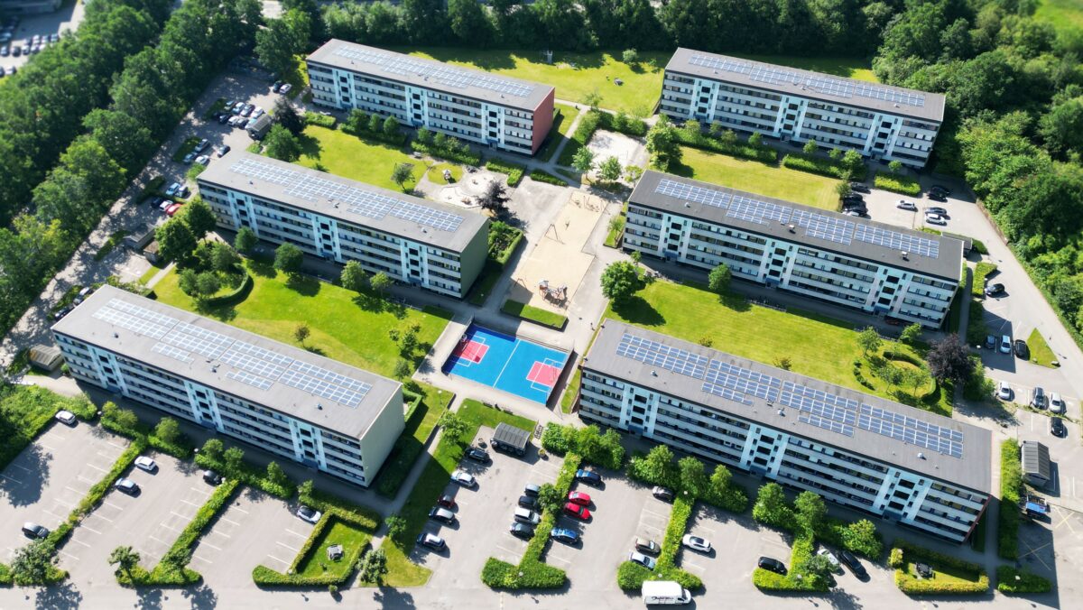 Luftfoto af de seks boligblokke i Vårkjærparken. Solceller på tagene, en sportsbane i midten i blå og rød, grønt græs mellem husene og parkeringspladserne
