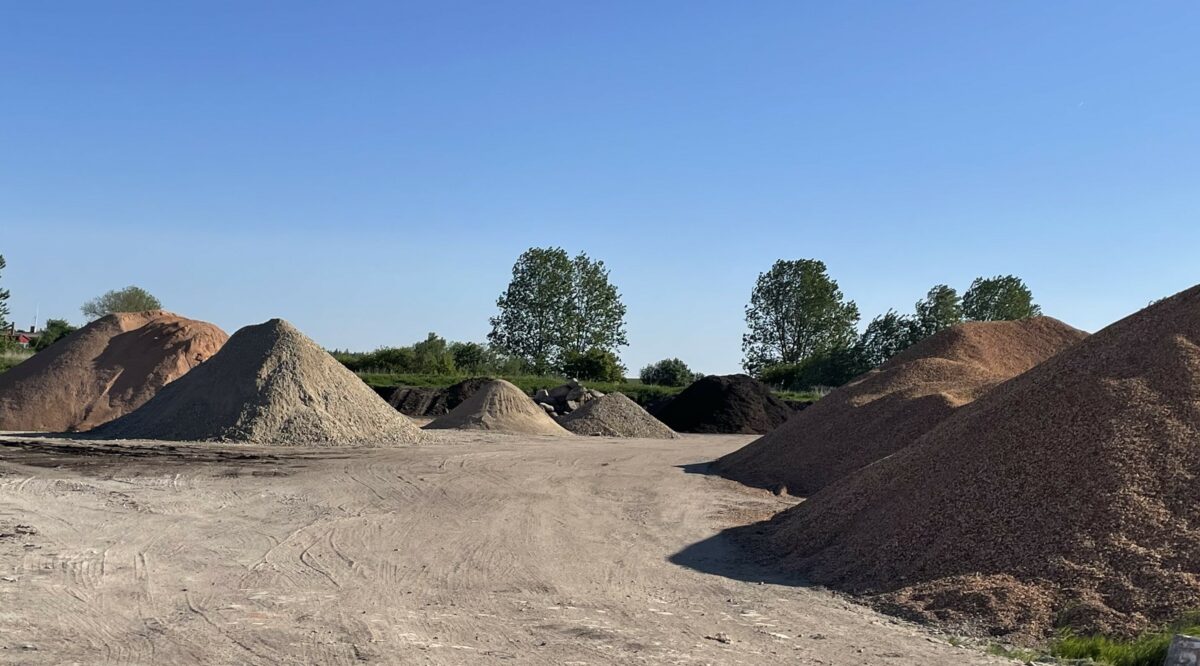 Store bunker af jord og grus i forskellige grå og brune nuancer. I baggrunden høje, grønne træer