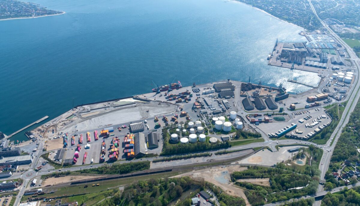 Luftfoto af Fredericia Havn med farverige og stablede containere, fragtskibe ved kaj og en område, der består af sand, der arbejdes på.