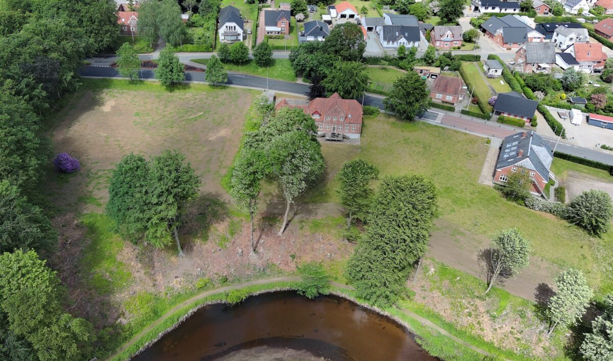 Luftfoto af en stor grøn plæne med en smule å med træer langs i bunden af billedet. Midt i billedet et rødligt hus med rødt tag. Bagest et villakvarter.