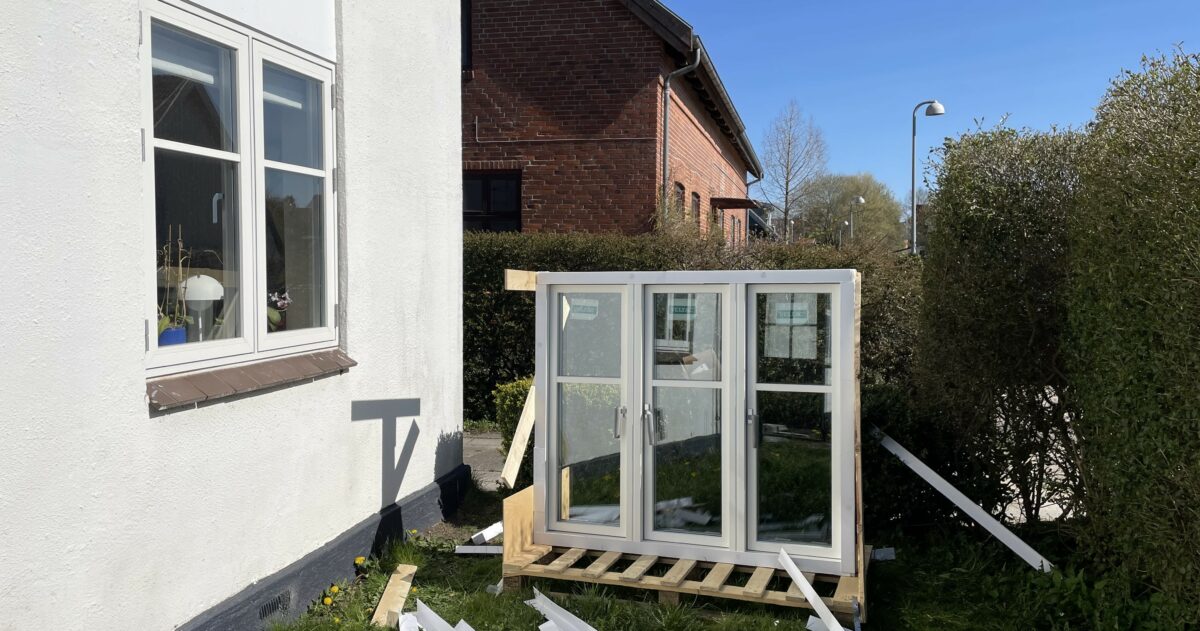 Foto af hvidt hus, der har fået nyt, moderne dannebrogsvindue. Et lignende, nyt vindue står på en træpalle på græsset. Til højre en hæk. I baggrunden et rødt hus.