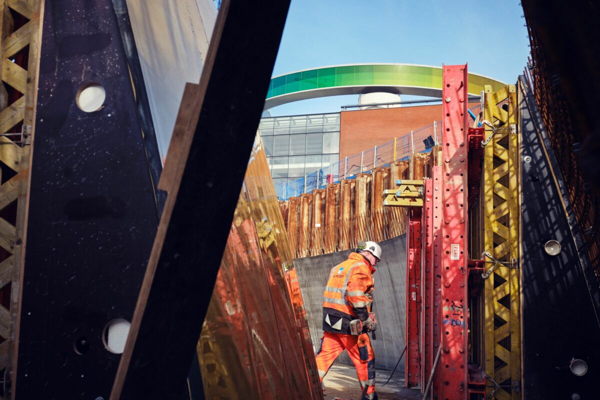 Mand i orange reflekstøj og hvid sikkerhedshjelm, går igennem byggeplads.