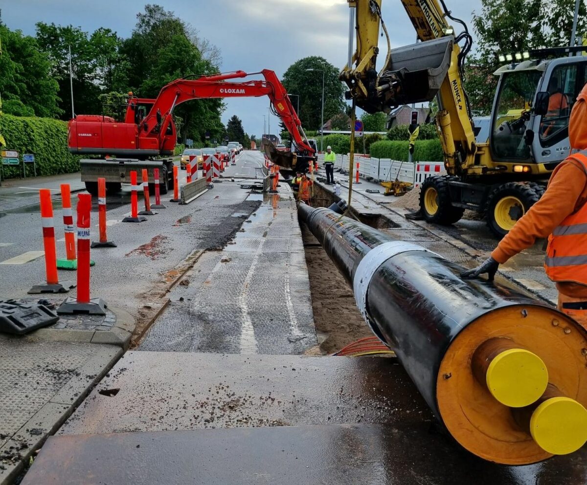 Foto af stort fjernvarmerør der er ved at blive lagt ned i en rende gravet i en asfaltvej. I baggrunden en rød kran. Ude til venstre står orange afspærringskegler