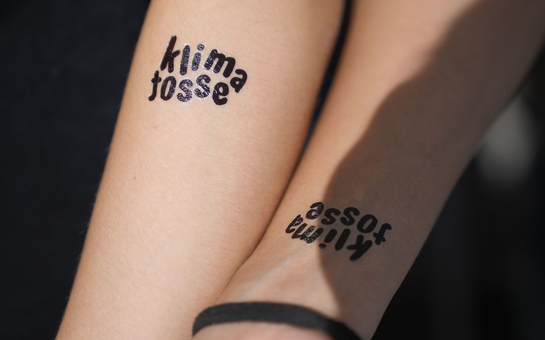 To slanke arme med hver sin klistertattooo, hvor der står klimatosse. Den ene håndled, hvor armen er lidt i skygge, er der et sort armbånd om