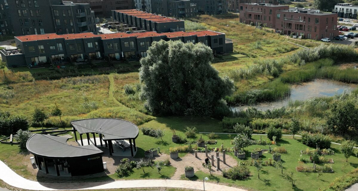 Luftfoto af et boligområde med røde blokke, lave rækkehuse, en sø og en rund, sort pavillon i træ, der er skåret bugtende over på midten. Byhaven står der med store bogstaver på væggen