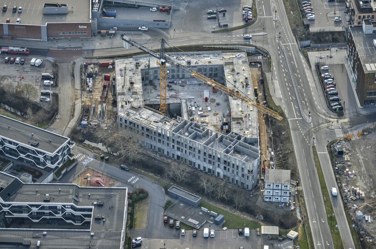 Luftfoto af de nye boligkompleks der er firkantet med med en skrå side. Store orange kraner troner over betonbyggeriet