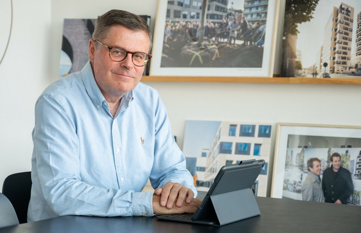 Søren Adamsen sidder ved et bord iført lyseblå skjorte. Foran en tablet. På væggen bag en samling fotos af mennesker og bygninger