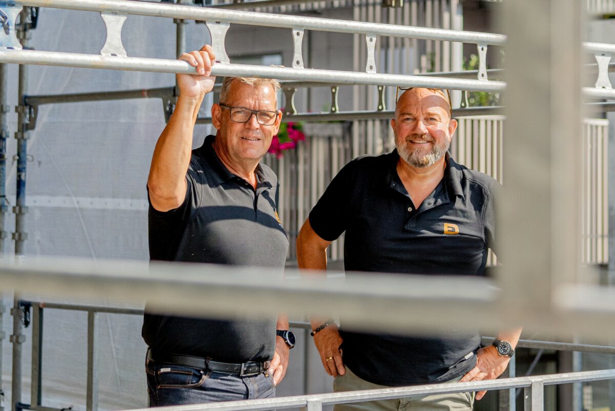 Steffen Povlsen og Karsten Andersen står side om side i matchende sorte poloer bag et stillads. Steffens højre hånd hviler på stilladset i ansigtshøjde.