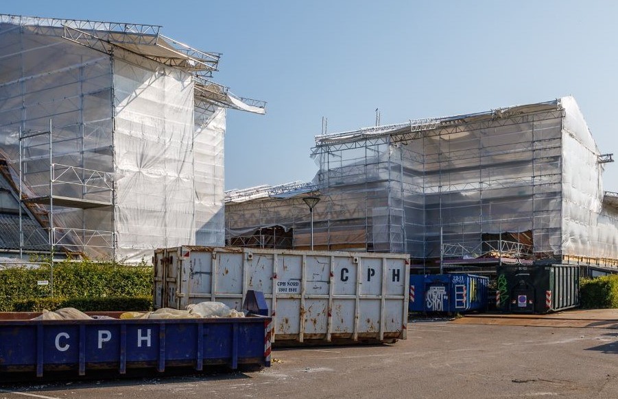 Foto af boligblokke pakket ind i stillads og hvide presenninger. Foan blokkene står to containere - en mørkeblå og en hvid. Himlen over blokkene er lyseblå