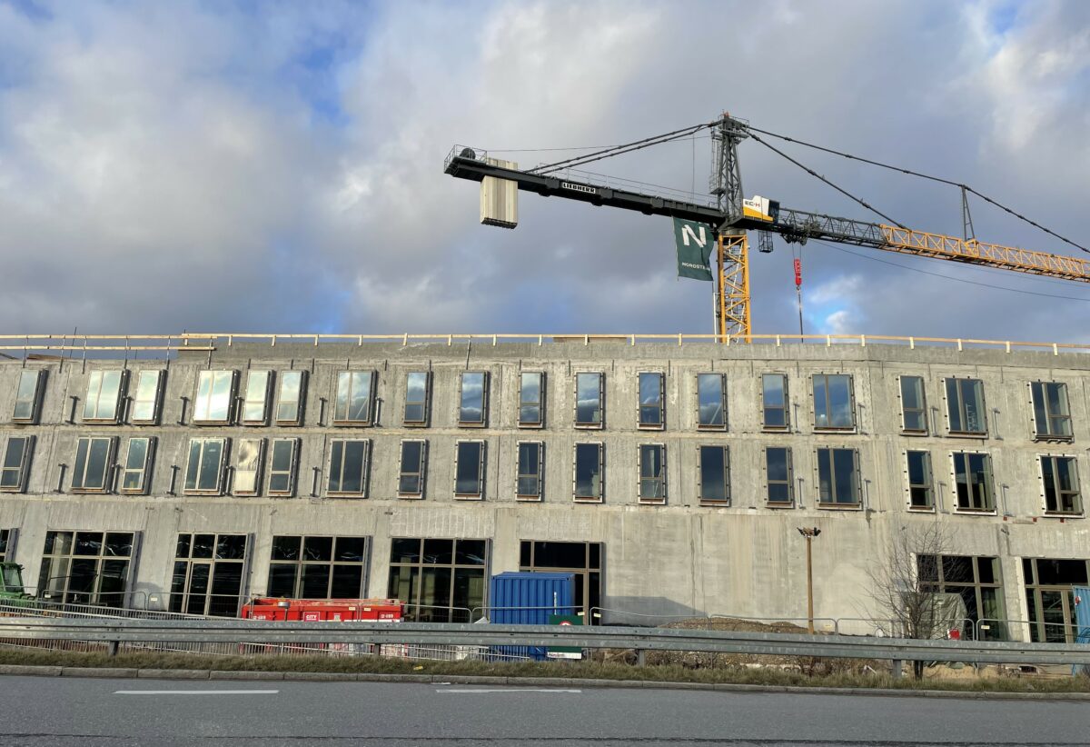 Foto af en lang stang beton i 3 etager, der skal blive til boliger. Der er ruder i vinduerne, men ellers står byggeriet råt. Bag byggeriet ses toppen af en gul kran