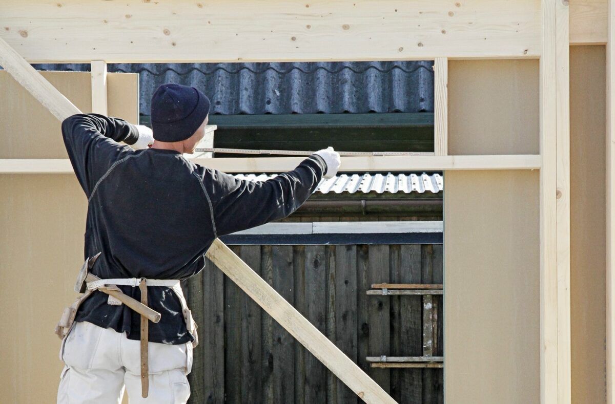 En person, set fra ryggen, iført hvide arbejdsbukser og sort trøje måler et stykke træ med en målestok.