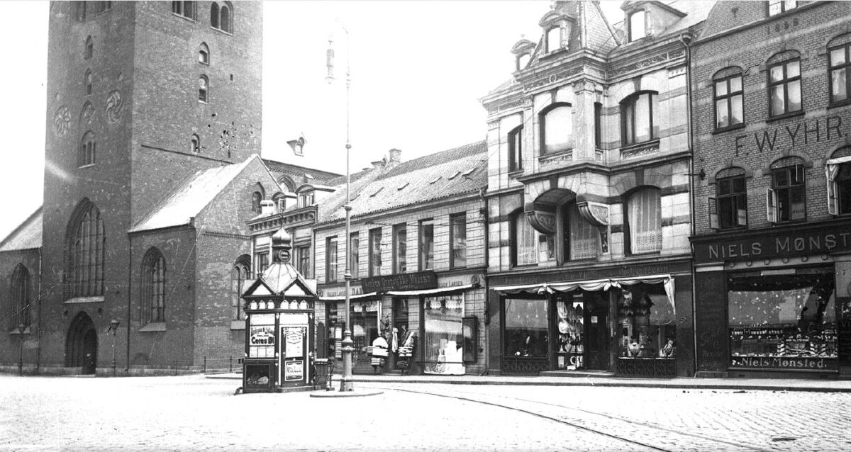 svart-hvitt bilde av den nedre delen av Århus domkirke - på torget kan du se en aviskiosk og flere butikker