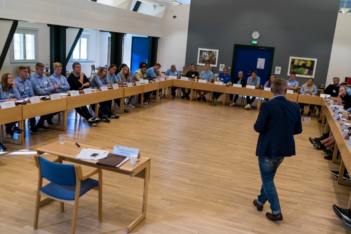 En hestesko med unge mennesker - de fleste i lyseblå skjorter - lytter til en mand i mørk jakke og cowboybukser, der står i midten. Han har et lille bord med en træstol med blåt stof til sin rådighed. Gulvet er lyst træ. Bagvæggen er mørkegrå. De andre vægge er hvide