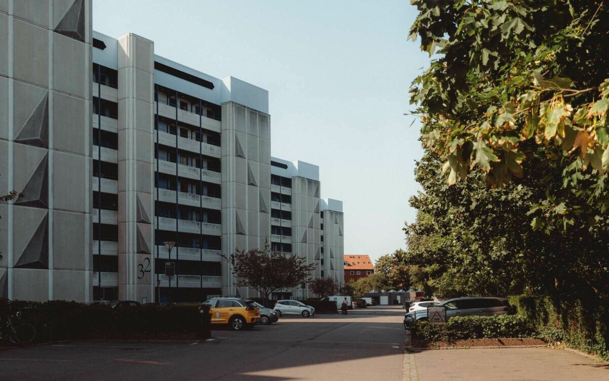 Foto af flere høje, lyse blokke. Der holder biler foran. Træer til højre på den anden side af indkørselsvejen