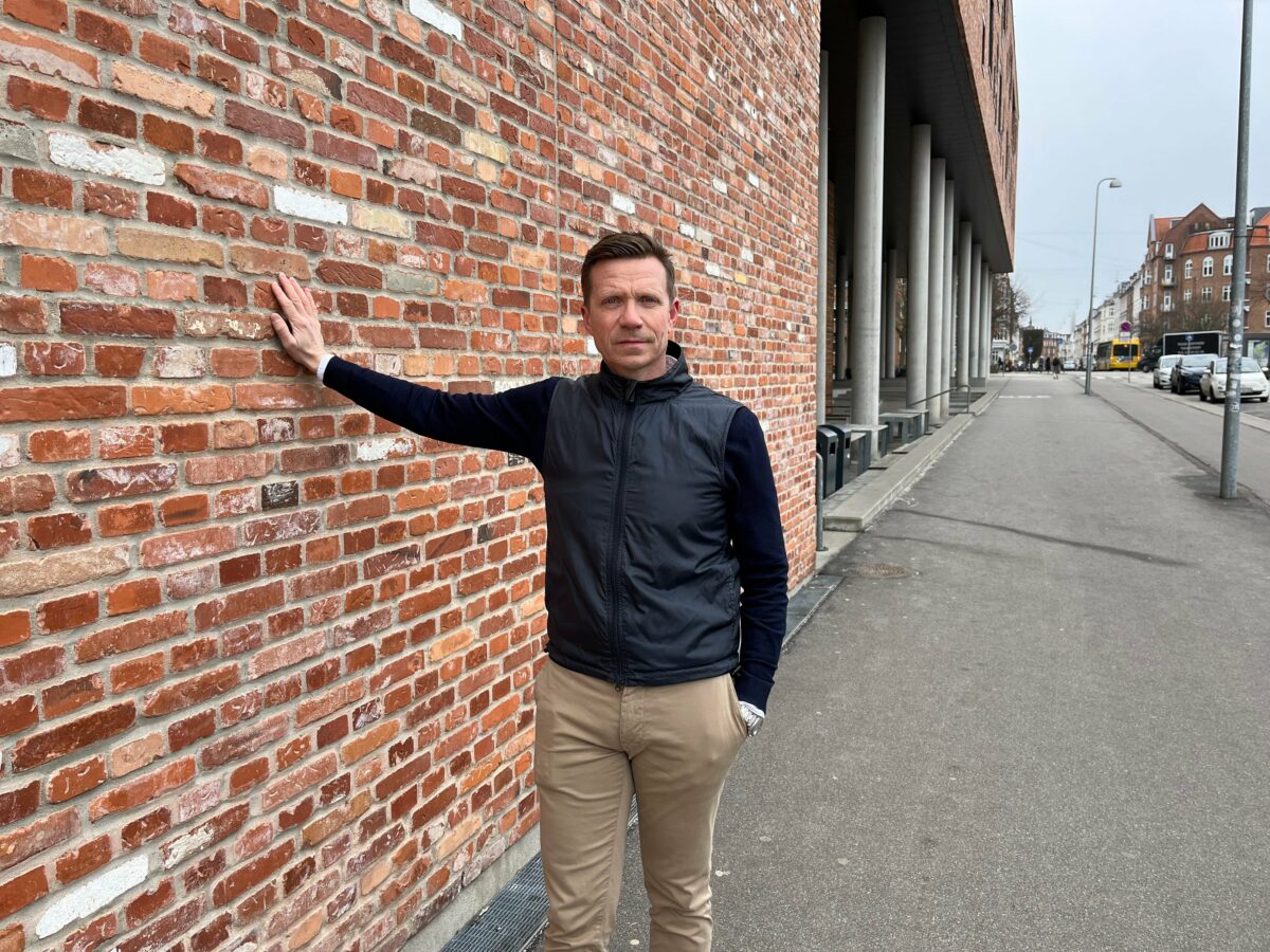 Jan Rhode i mørk trøje og mørk dunvest står ved en mur af genbrugte teglsten. Han har lagt sin højre hånd flat mod muren. Armen er strakt. Han har beige bukser på