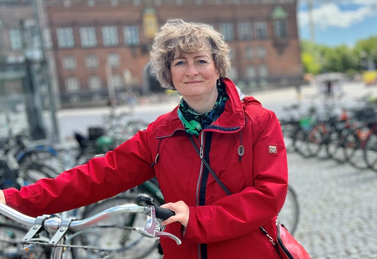 Line Barfod står med sin cykel foran Københavns Rådhus. hun har rød jakke på, rød taske og der ligger en rød cykelhjelm i cykelkurven foran på cyklen