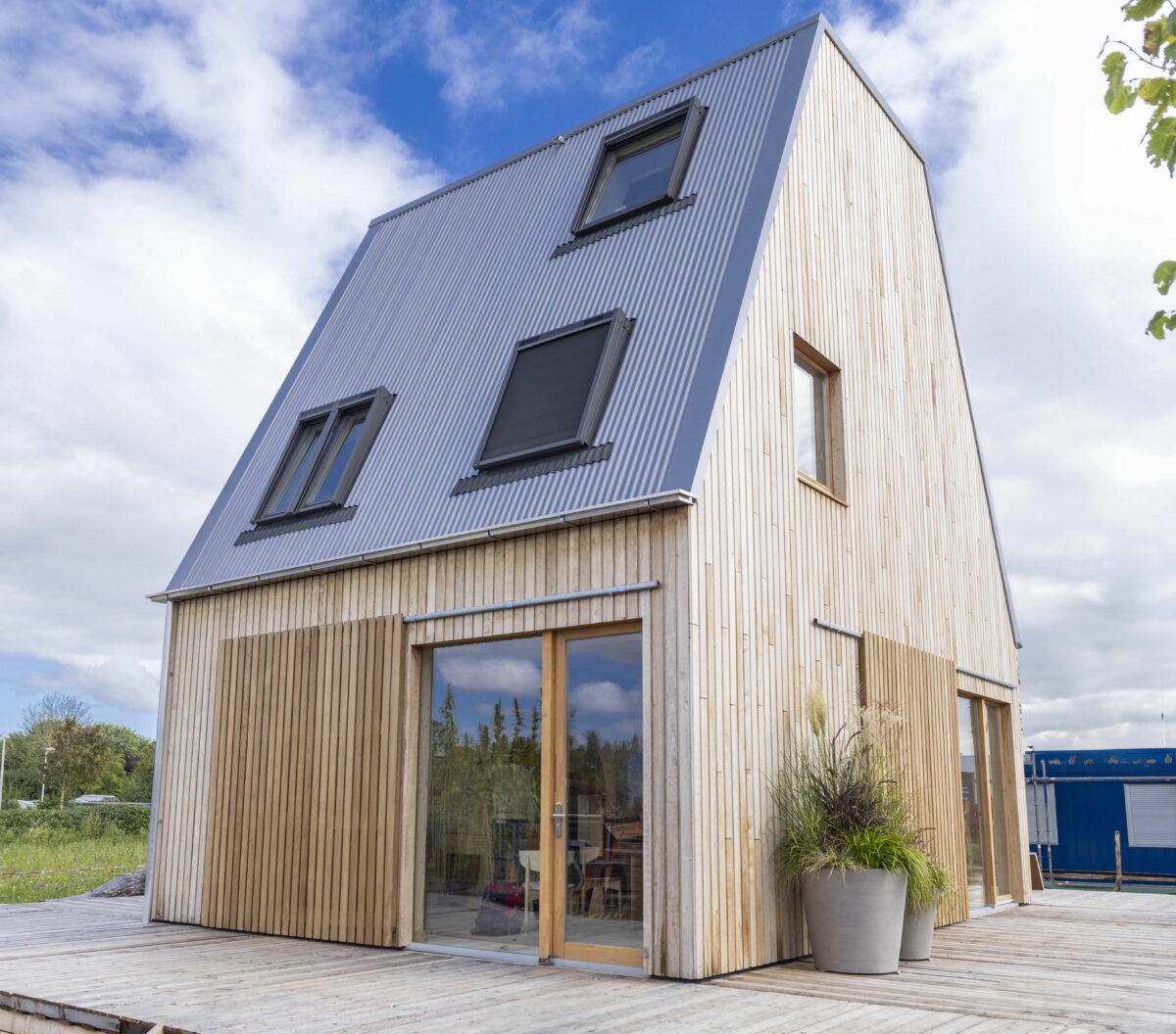 Foto af træhus med meget høj tagrejsning, tre vinduer i tagfladen, et lille i tagetes gavl. På facaden flere store vinduer på de to flader der ses. Der står en krukke med en plante op af huset. til højre ses en smule af et træ