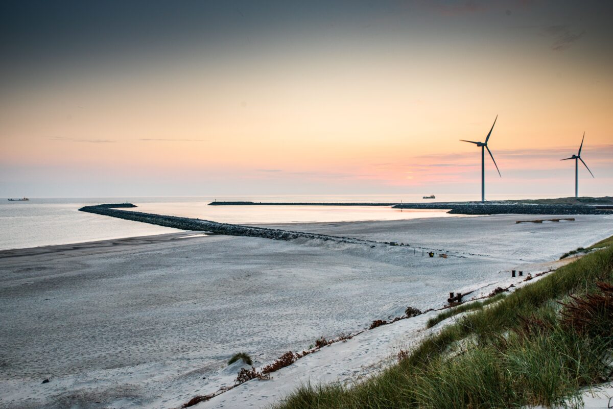 Dronebillede af en strand med solnedgang og havvindmøller i horisonten