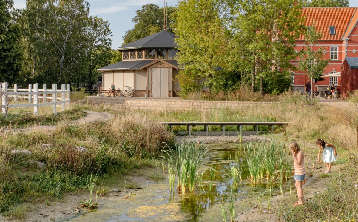 Foto af to piger der leger ved en lille sø. til venstre ses hegnet til en hestefold. I baggrunden et ældre, rødt hus. en cykelrytter sidder på en bænk foran en kirkelignende, men lav bygning med sort tag