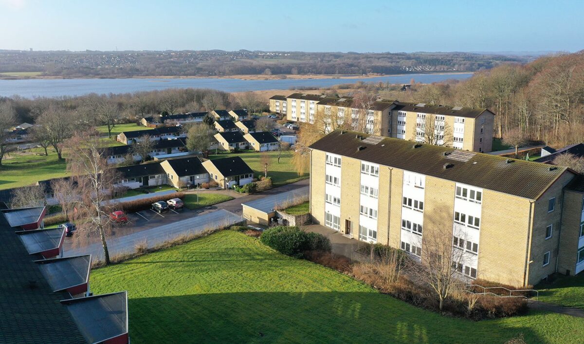 Luftfoto af boligområde med både gule etageboliger og hvide huse med sorte tage. I baggrunden ses en stor sø