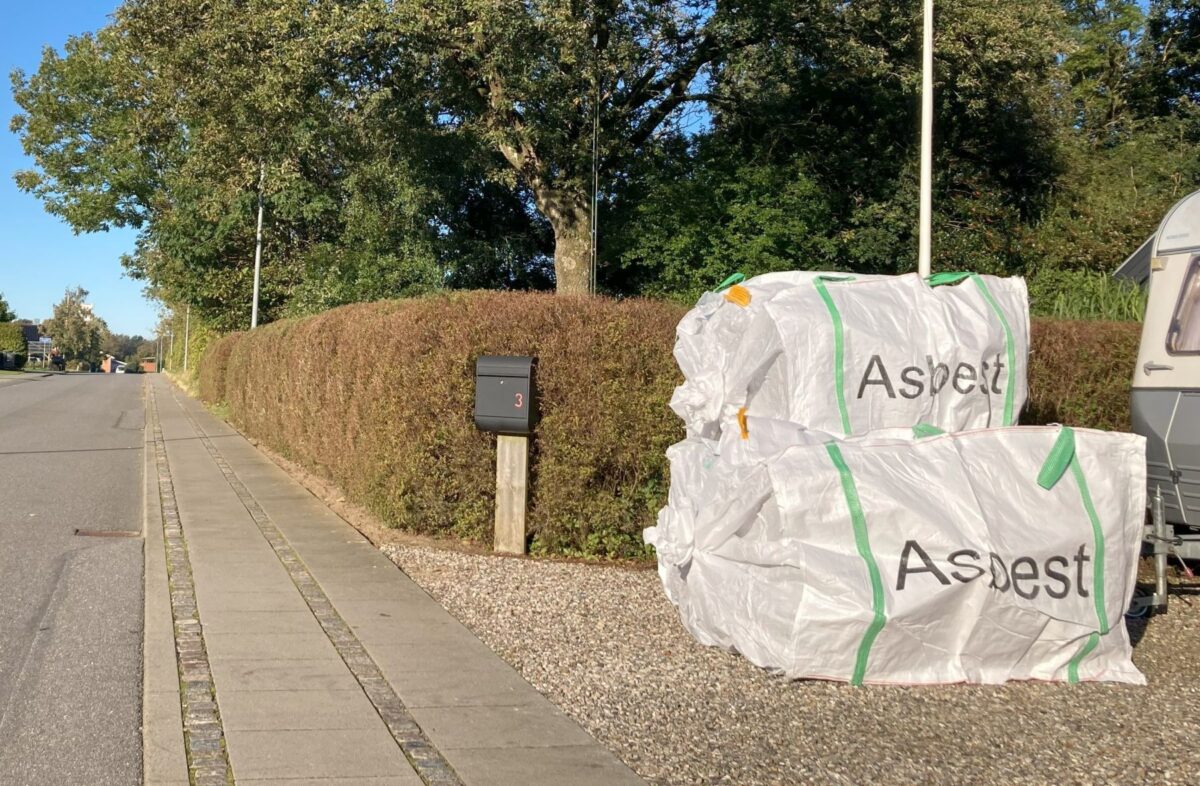 Foto af store hvide bigbags med påskriften Asbest, der står i perlegruset ved siden af en sort postkasse på en træpæl