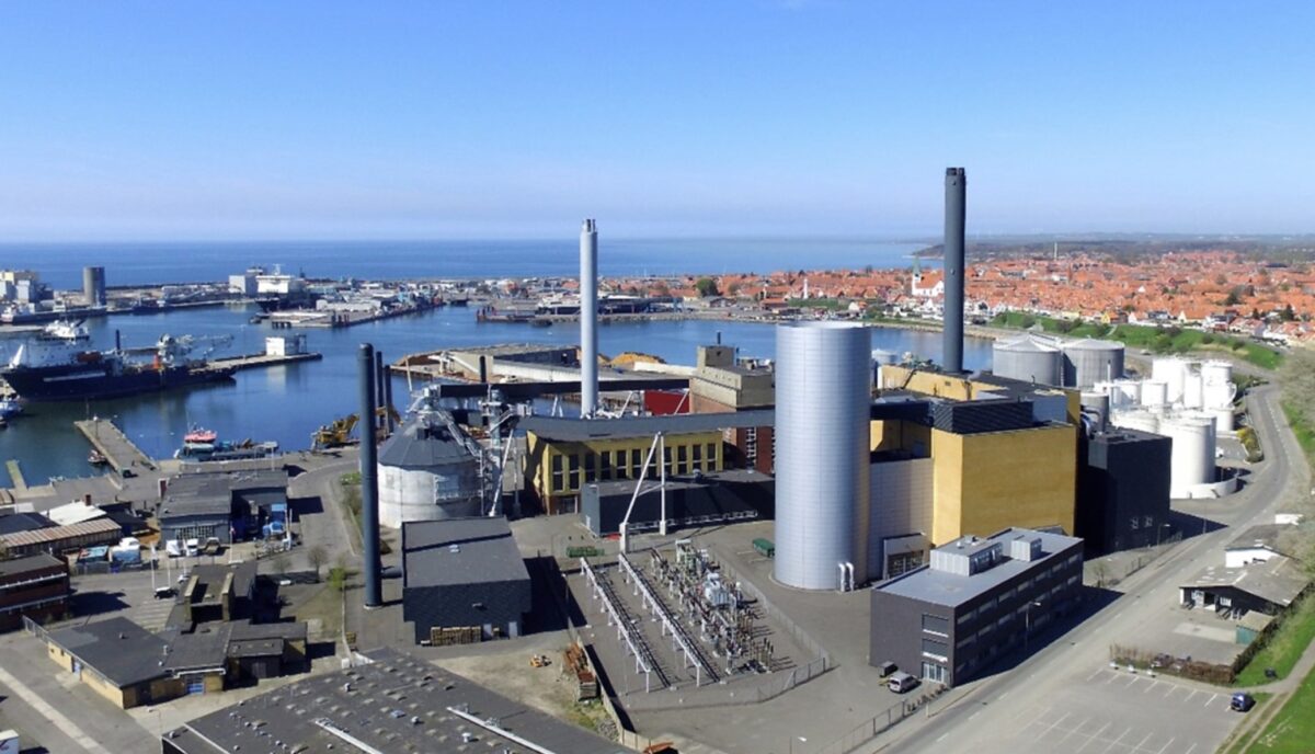 Overbliksfoto af et varmeværk med siloer og skorstene. I baggrunden vand og en havn