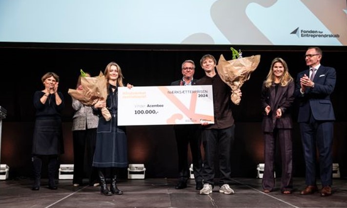 To unge mennesker holder en stor check med 100.000 kr. skrevet på. De holder begge en stor buket blomster i den hånd, der ikke holder papchecken