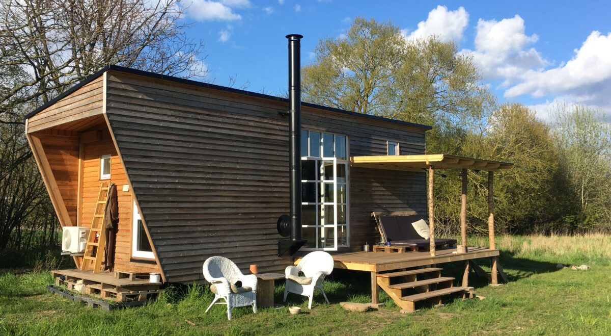 Tiny house bygget i træ med sort skorsten, trappe op til boligens indgang hvor der også er en overdækket terrasse med siddeplads med hynder. Der står hvide havemøbler på græsset foran boligern