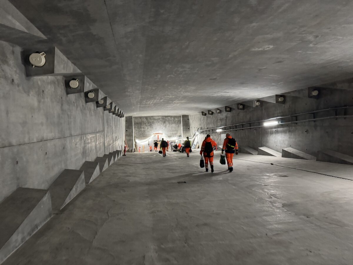 Foto fra innsiden av broen - en bred betonggang der en gruppe mennesker i oransje og mørkeblå klær beveger seg bort fra kameraet