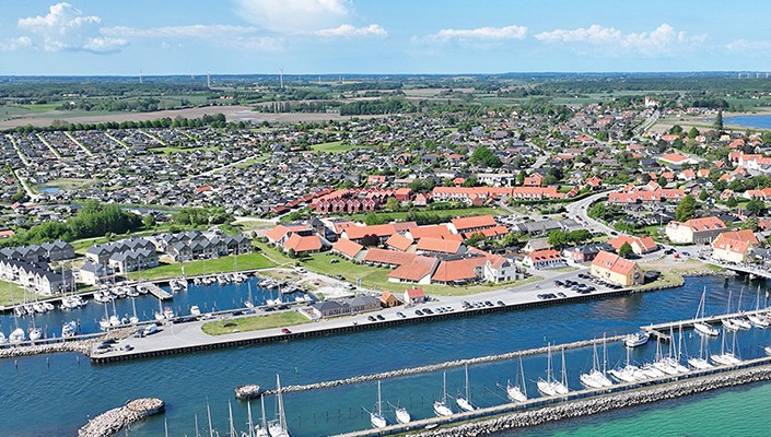 Dronefoto af Karrebæksminde med lystbådehavnen i forgrunden. På den anden side af havnens vand ligger en stor samling huse med store, røde tagflader.