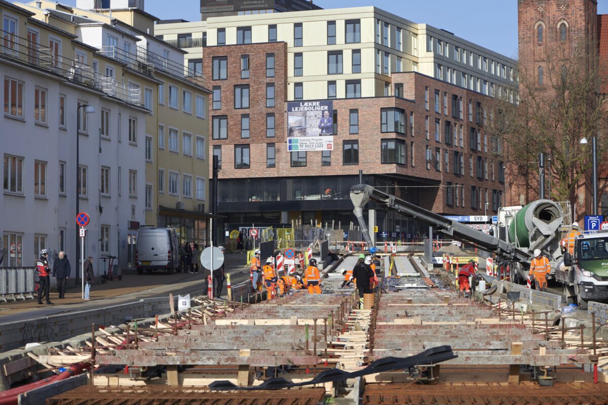 Maskiner og mennesker færdes på byggeplads, midt inde i byen.