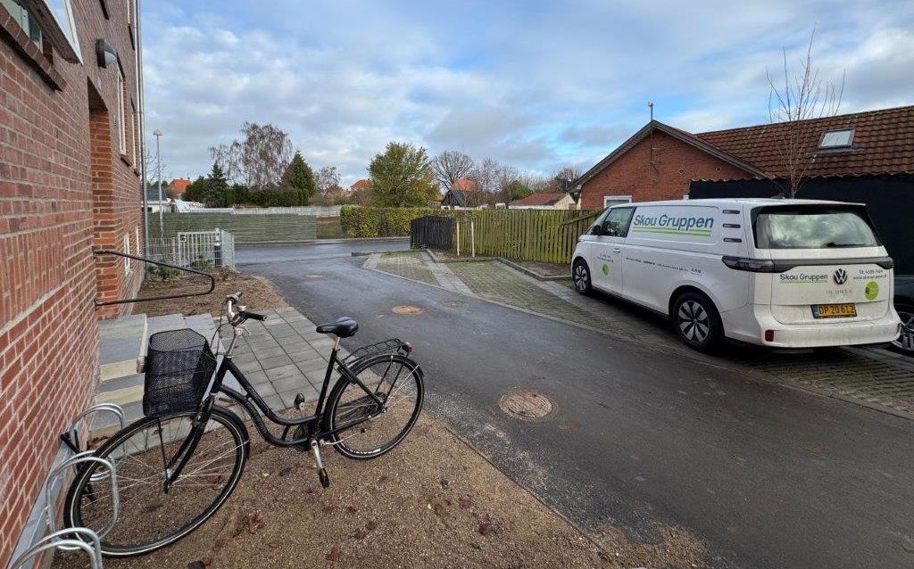 Foro af rød boligblok til venstre. Foran holder en sort cykel parkeret i et stålgråt cykelstativ. På den anden side af en smal vej holder en hvid varevogn