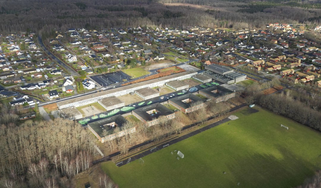 Luftfoto af den nuværende skole, der er opbygget i fire firkanter med en åben midte samt fem øvrige, kvadratiske bygninger bygget sammen med en lang, hvid bygningsstang. I forgrunden en fodboldbane