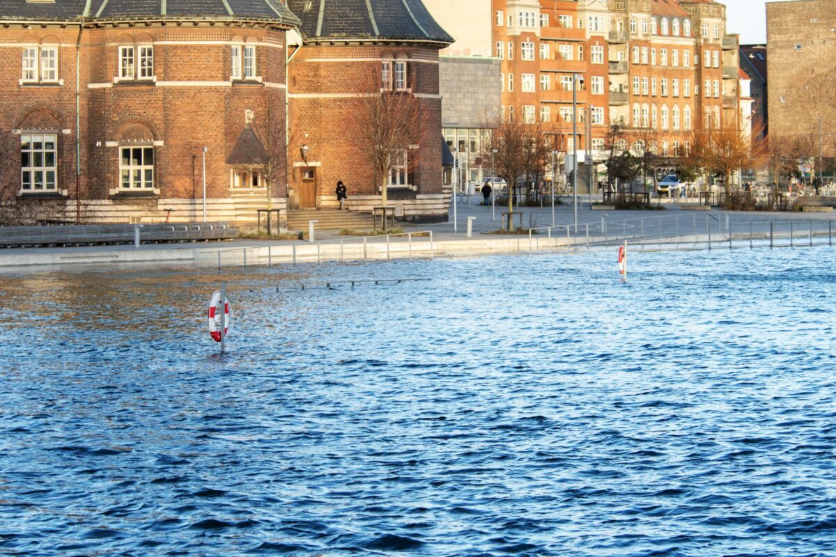 Foto af havvand, der er gået over sine breder. Vandet når op til en vej. På den anden side af vejen ligger nogle ældre, røde murstensbygninger