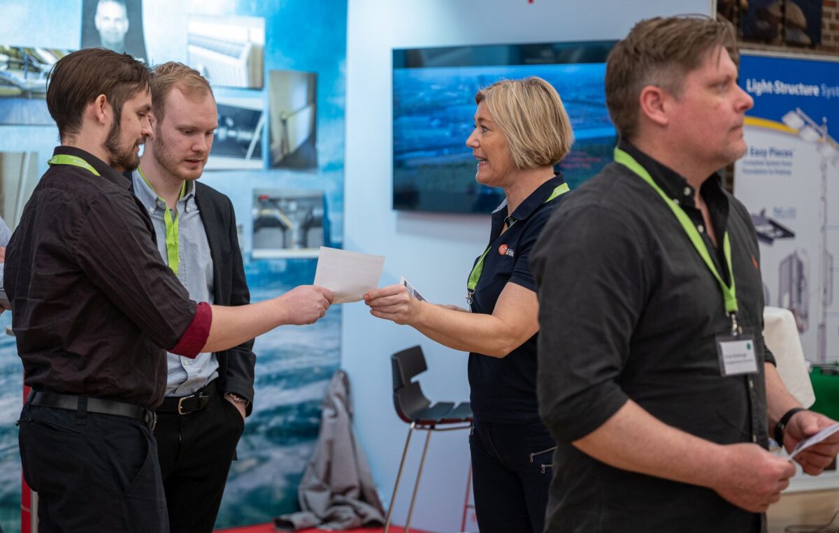 Foto fra en konference. En kvinde på en stand giver et stykke papir til en mand. To andre mænd med grønne  nøglesnore om halsen er også i billedet