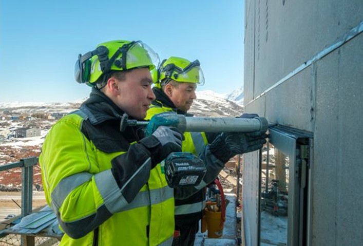 To mænd i gult og sort  arbejdstøj og gule hjelme fuger omkring et vindue. I baggrunden ses soloplyst fjeld med sne