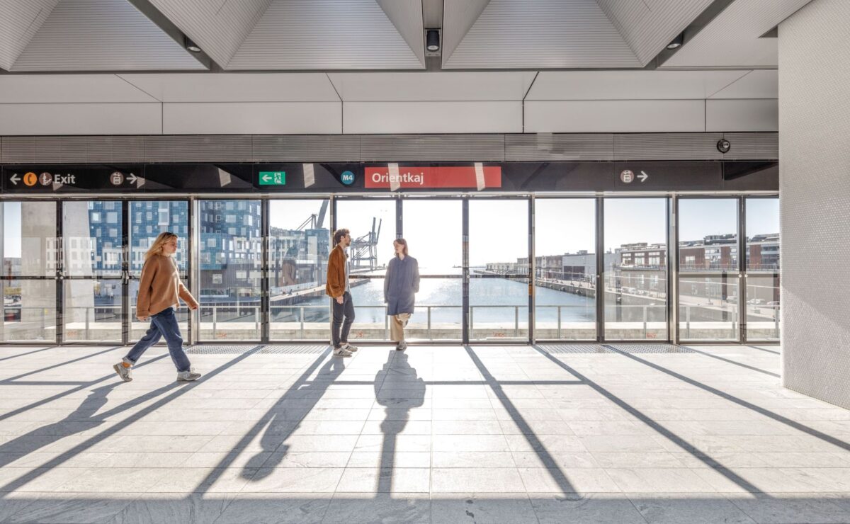 Foto af en metrostation med vandkig ud gennem store ruder. Et par læner sig op af ruderne. En kvinde går forbi