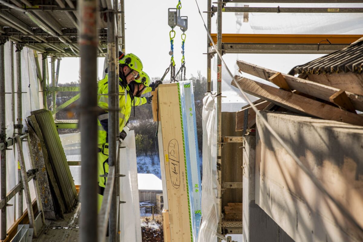 Nye træfacader kranes op på ydersiden af en bygning. To mænd i skriggult arbejdstøj tager imod