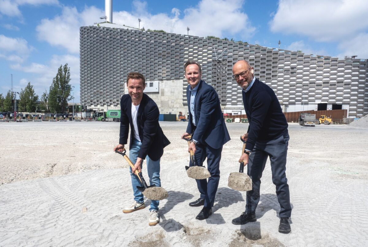 Tre mænd med hver deres spade står på en grusplads foran Amager Bakke