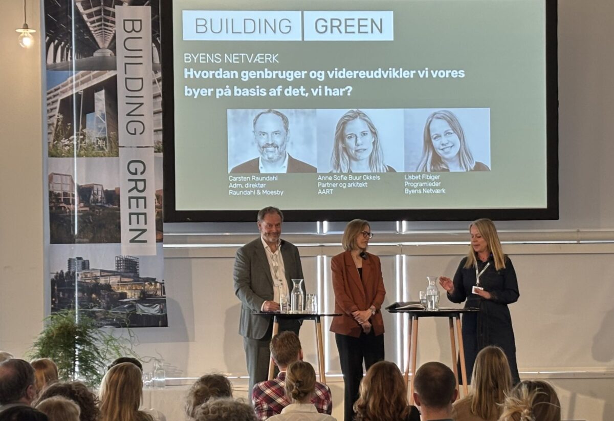 Foto fra en scene på Building Green med tre stående mennesker, der taler sammen foran et siddende publikum
