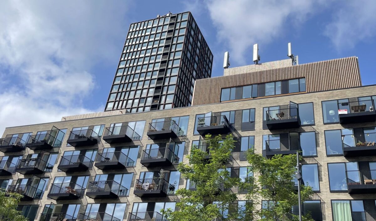 Foto af en lavere bygning med lejligheder med sorte altaner og træer foran. Ovenover troner et langt højere højhus