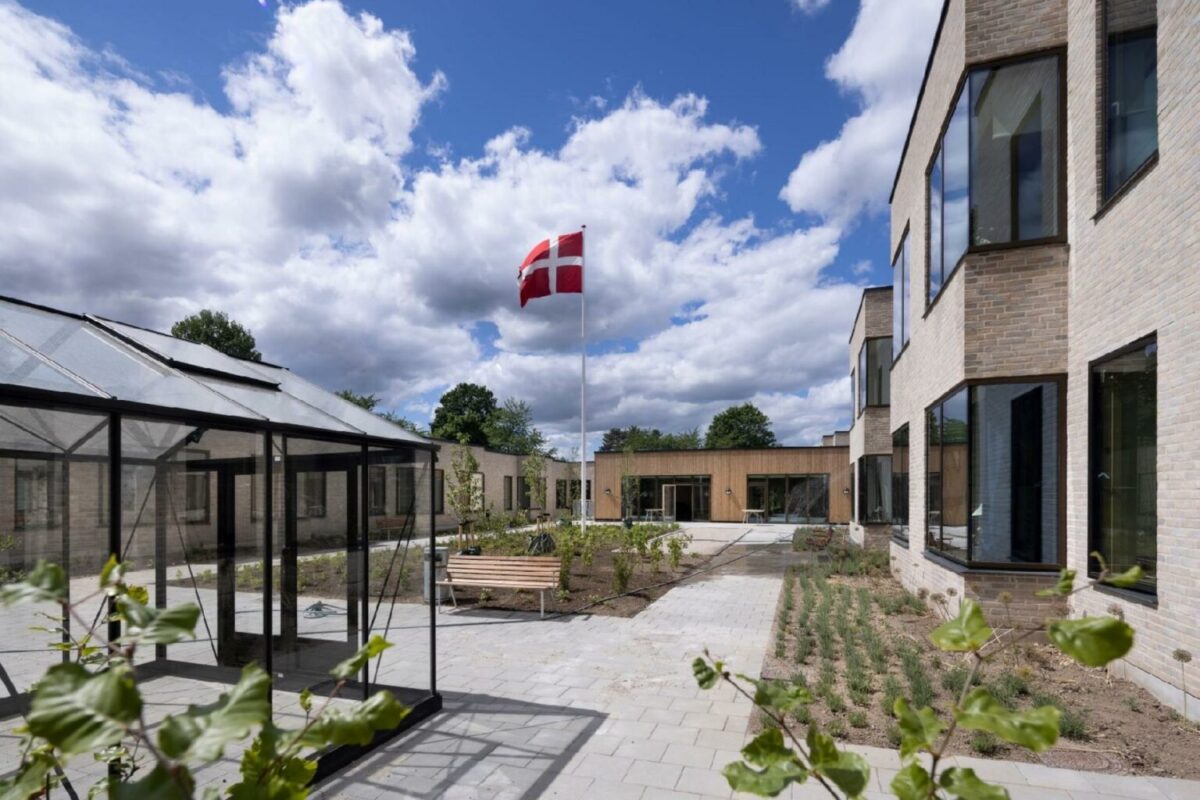 Foto af gårdrum mellem lyse bygninger. I forgrunden et drivhus