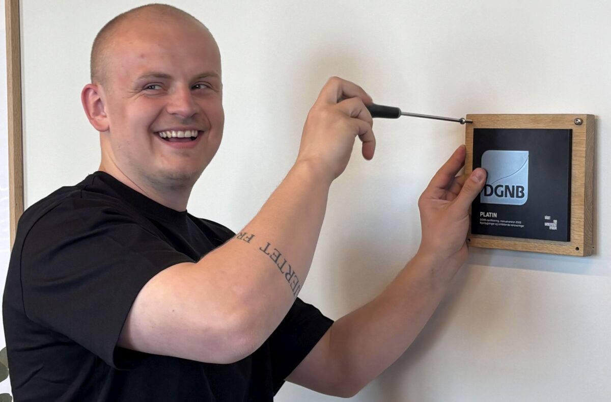 En smilende mand i sort t-shirt i færd med en skrue en plakette i træ og sort fast på væggen