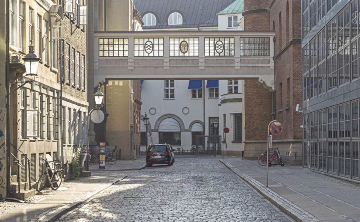 Foto ned langs en brostensbelagt gade. I enden af gaden ses en gangbro der forbinder bygningerne på hver sin side af gaden