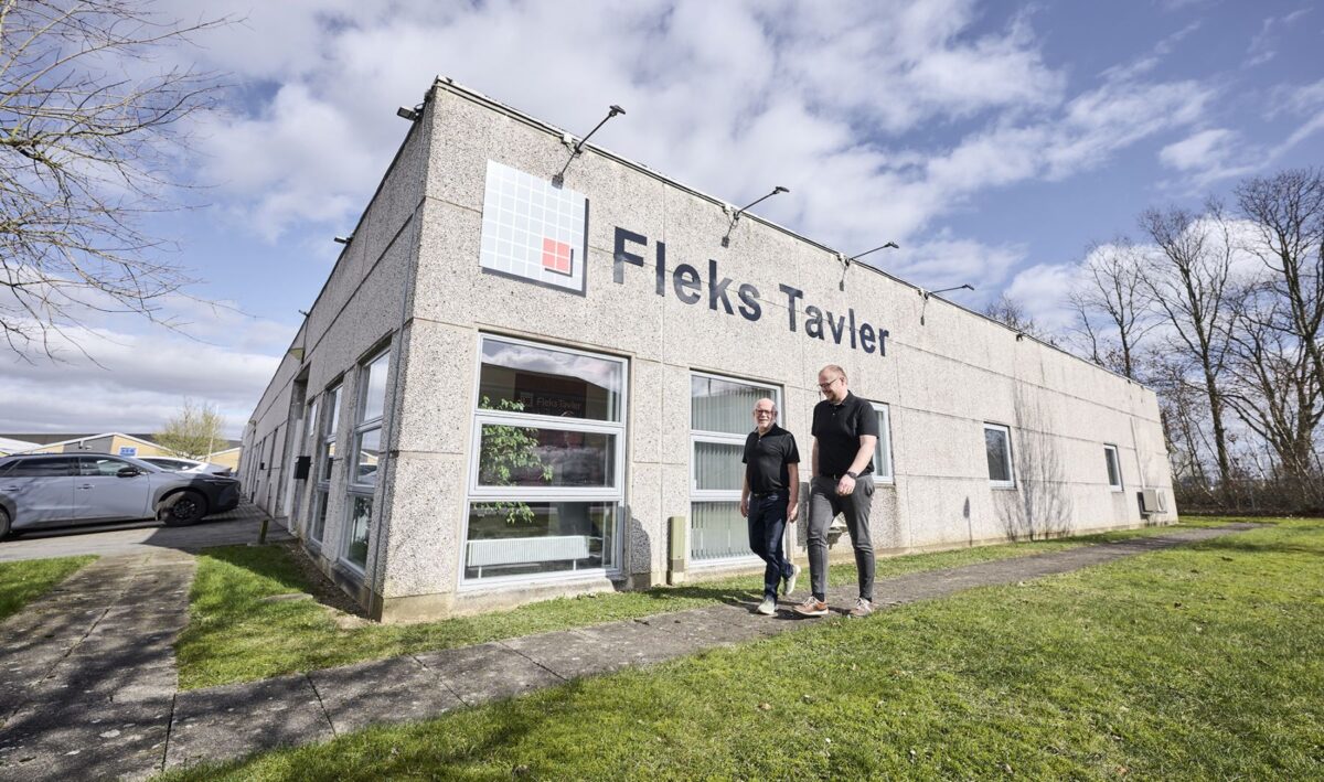 Foto af to mænd i sorte poloer, der går rundt om en grå industribygning med navnet Fleks Tavler på facaden