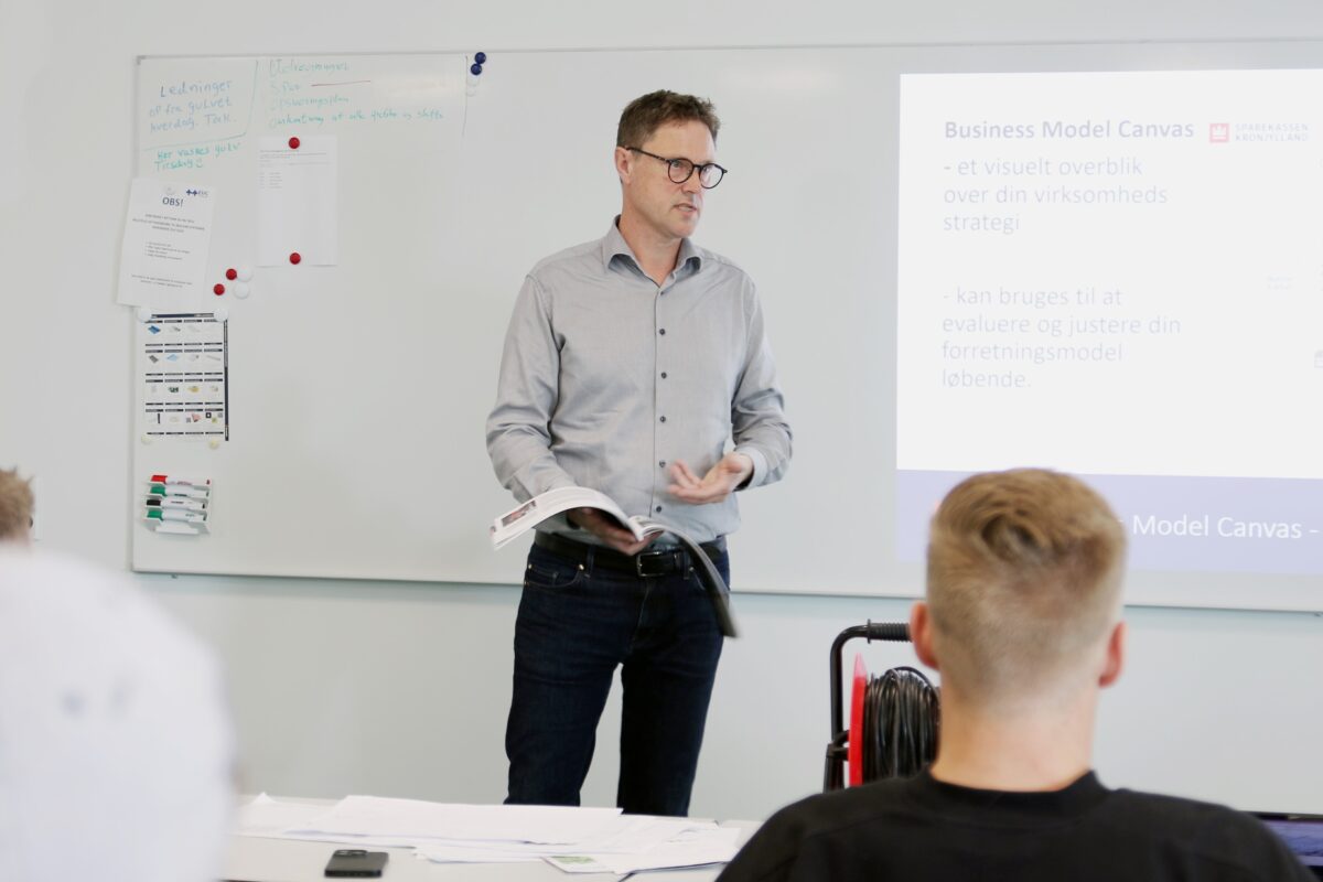 En mand står foran et whiteboard med en bog i hånden og taler.