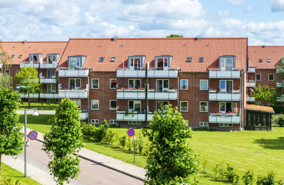 Foto af en boligforening med en grøn græsplæne og høje træer foran. Bygningen er rød med hvide altaner